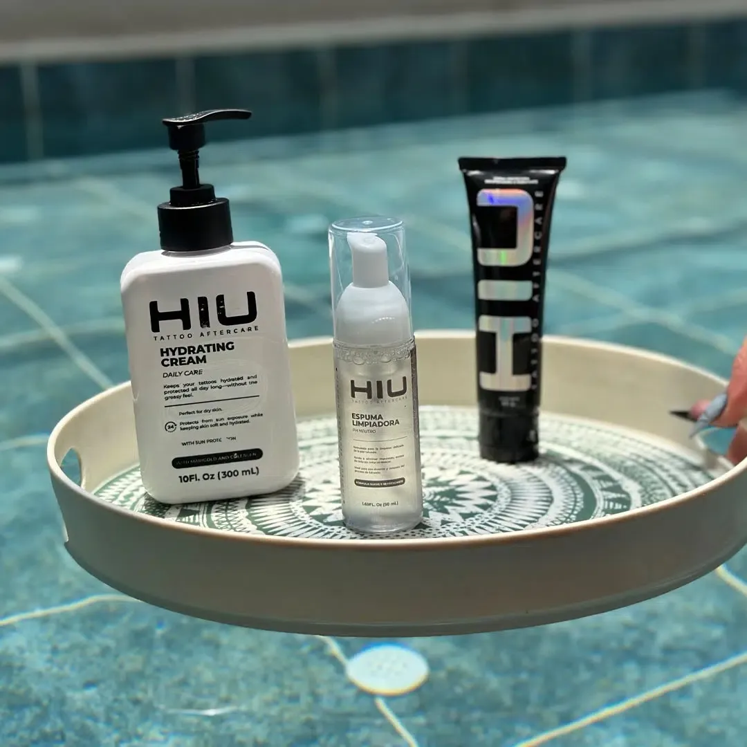 Trayedor con tres productos de cuidado post-tatuaje: crema hidratante en bomba, loción en spray y tubo de pomada, sobre fondo de piscina.