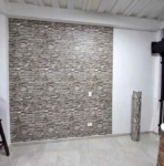 Papel tapiz adhesivo PVC en textura de piedra gris, instalado en pared interior con enchufes visibles y rodillo de muestra al lado.
