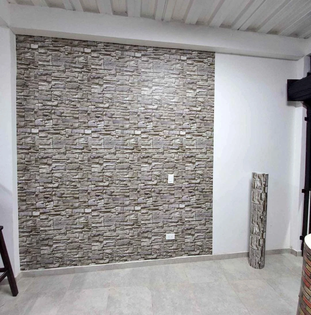 Papel tapiz adhesivo PVC en textura de piedra gris, instalado en pared interior con enchufes visibles y rodillo de muestra al lado.