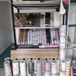 Rollo papel tapiz adhesivo PVC con diseños variados, exhibido en estantería y rollos apilados en el suelo.