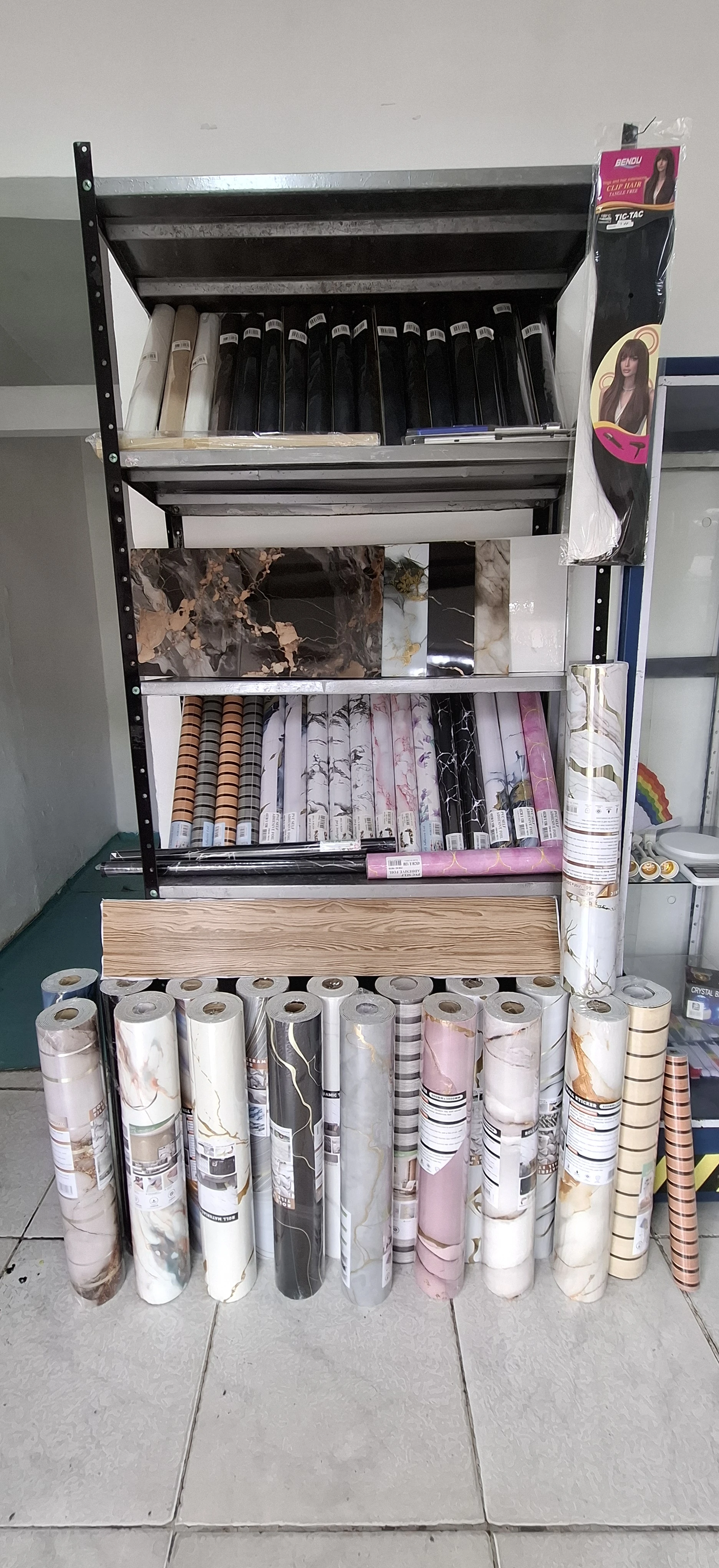 Rollo papel tapiz adhesivo PVC con diseños variados, exhibido en estantería y rollos apilados en el suelo.