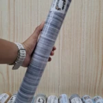 Rollo de papel tapiz adhesivo PVC con patrón gris y blanco, enrollado y sostenido en mano, mostrando etiqueta y textura visible.