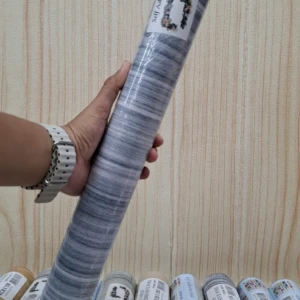 Rollo de papel tapiz adhesivo PVC con patrón gris y blanco, enrollado y sostenido en mano, mostrando etiqueta y textura visible.