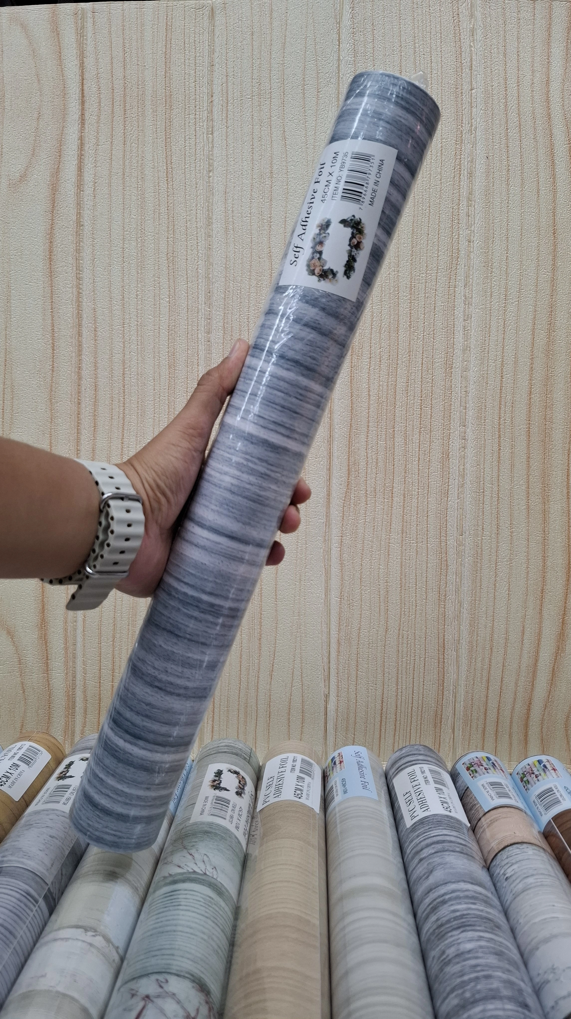 Rollo de papel tapiz adhesivo PVC con patrón gris y blanco, enrollado y sostenido en mano, mostrando etiqueta y textura visible.