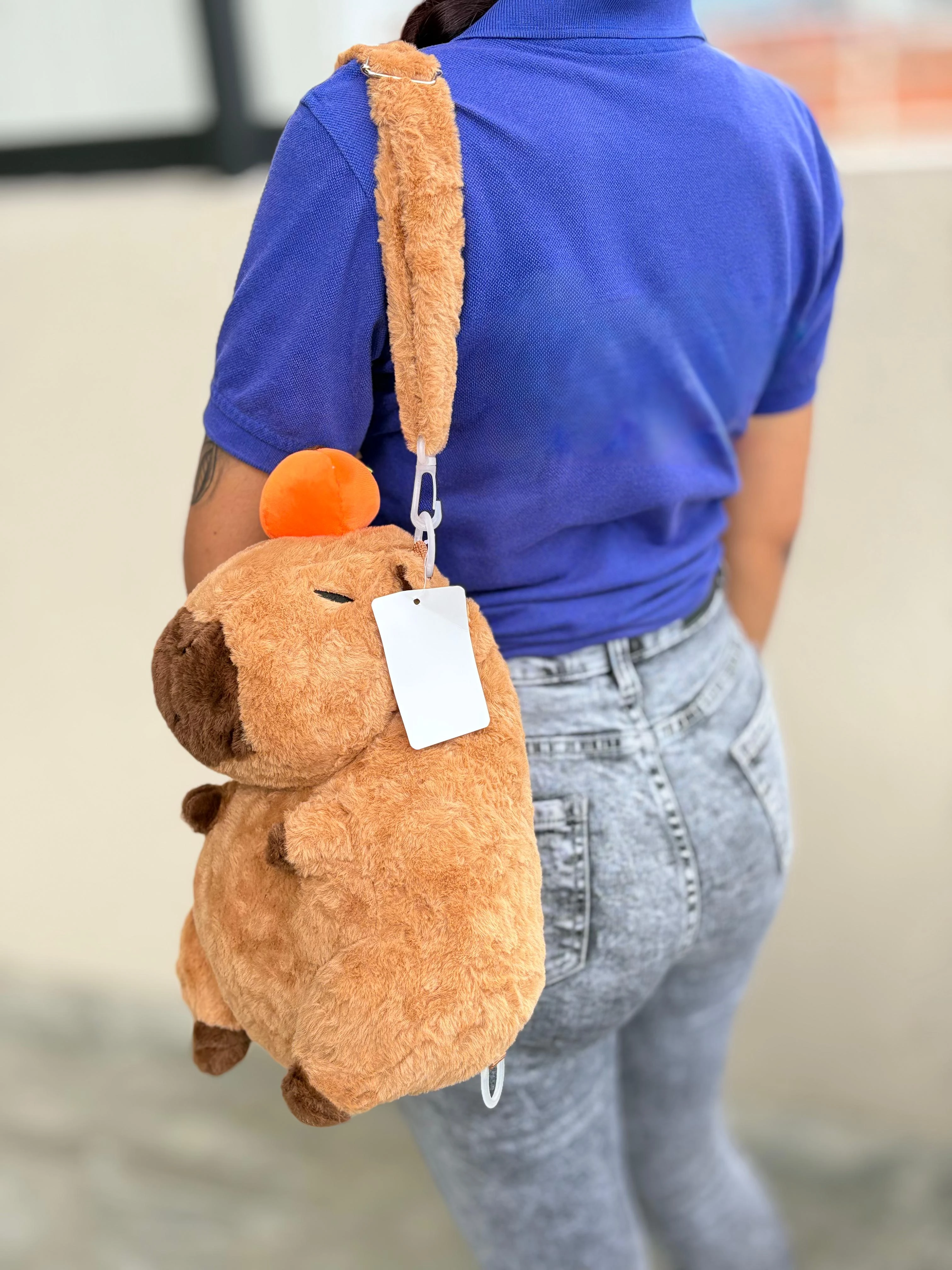 Mochila de peluche capibara marrón con orejas naranjas, usada como bolso de hombro por persona con camiseta azul y jeans.