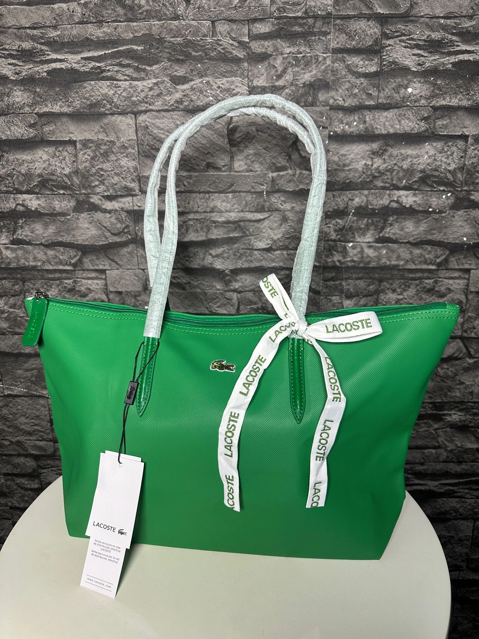 Bolso tote verde brillante de Lacoste con asas trenadas en tono claro, detalles en blanco y etiqueta colgante visible sobre fondo de pared de ladrillo.