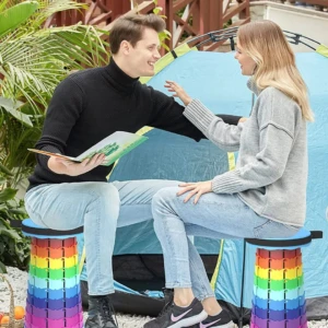 Dos taburetes plegables con base antideslizante, color arcoíris, en uso al aire libre por una pareja sentada frente a una carpa azul.
