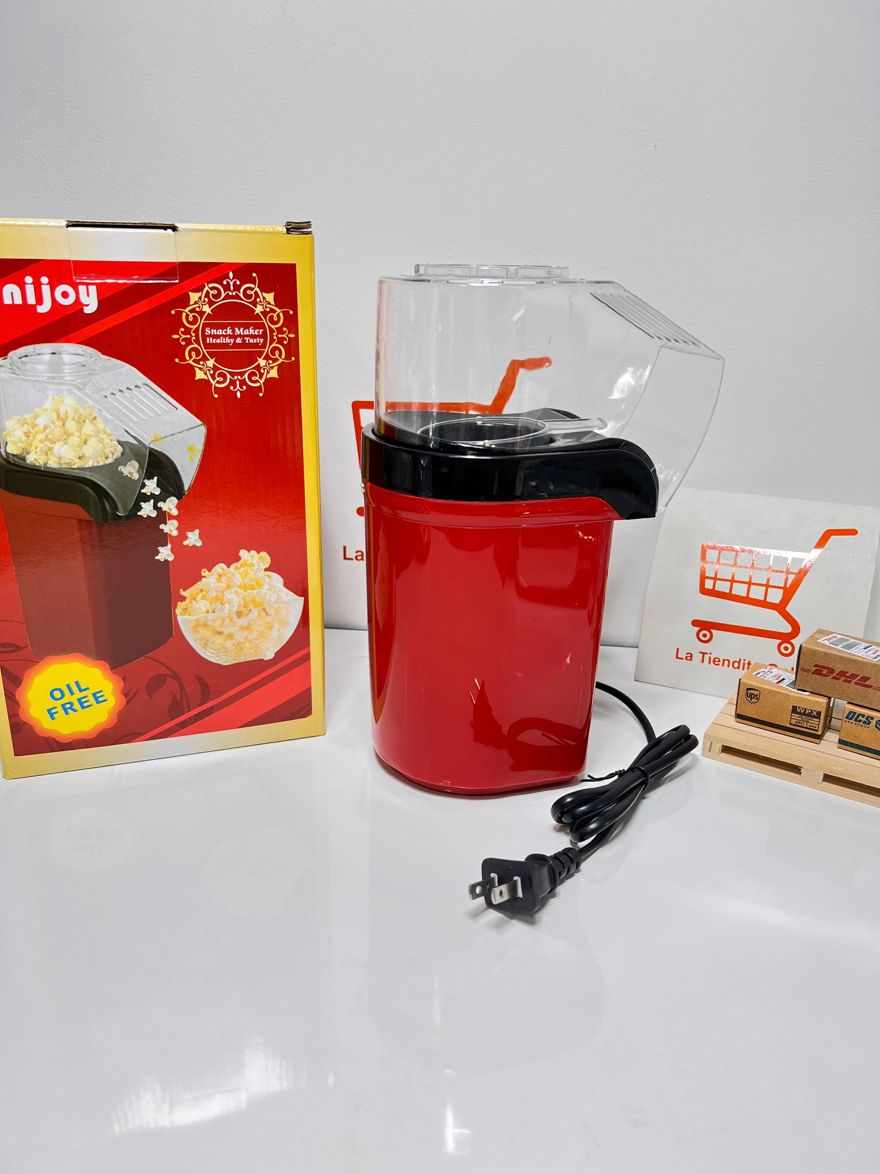 Crispetera eléctrica roja con tapa transparente, cable negro y caja de cartón al lado.