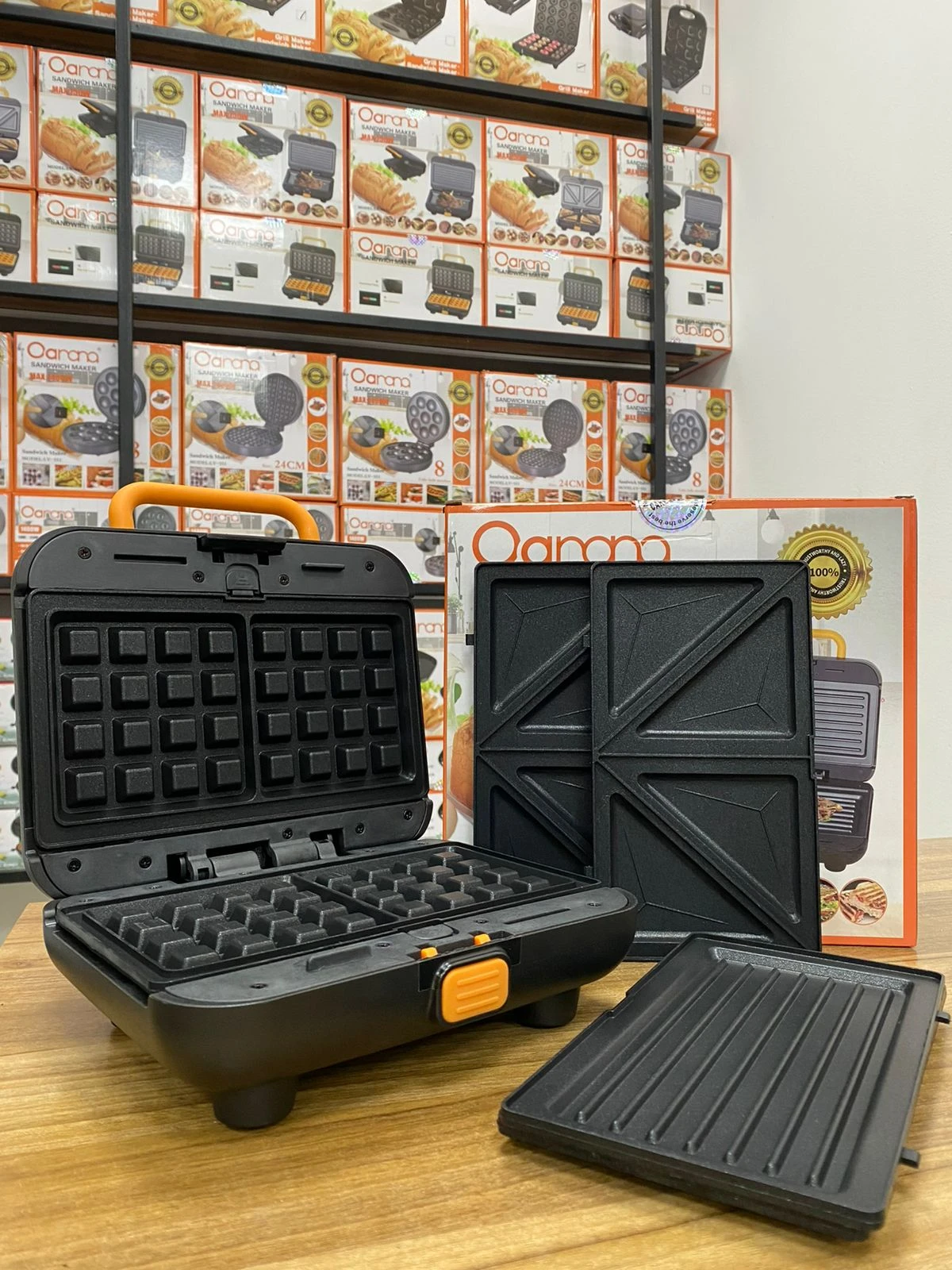 Máquina 3 en 1 para sándwiches, waffles y parrilla, color negro, abierta sobre mesa de madera, con bandejas intercambiables visibles y caja de producto al fondo.