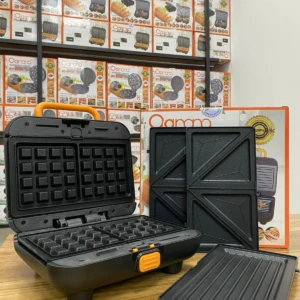 Máquina 3 en 1 para sándwiches, waffles y parrilla, color negro, abierta sobre mesa con accesorios desmontados y caja de marca Ocampo al fondo.