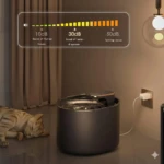 Bebedero automático para gatos en color gris oscuro, con agua en movimiento y gato durmiendo al lado. Incluye indicador de decibelios y está conectado a la pared.