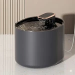Bebedero automático para mascotas en color gris oscuro, con tapa negra y dispensador de agua en funcionamiento sobre superficie blanca.
