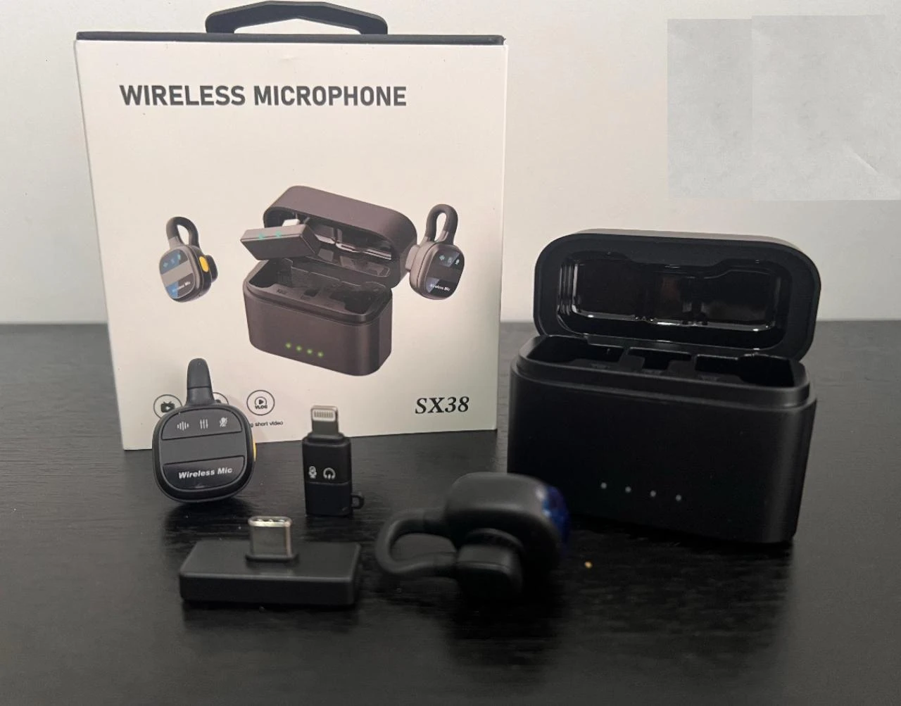 Caja de producto inalámbrico modelo SX38 con estuche de carga negro abierto, micrófono y accesorios dispuestos sobre superficie oscura.