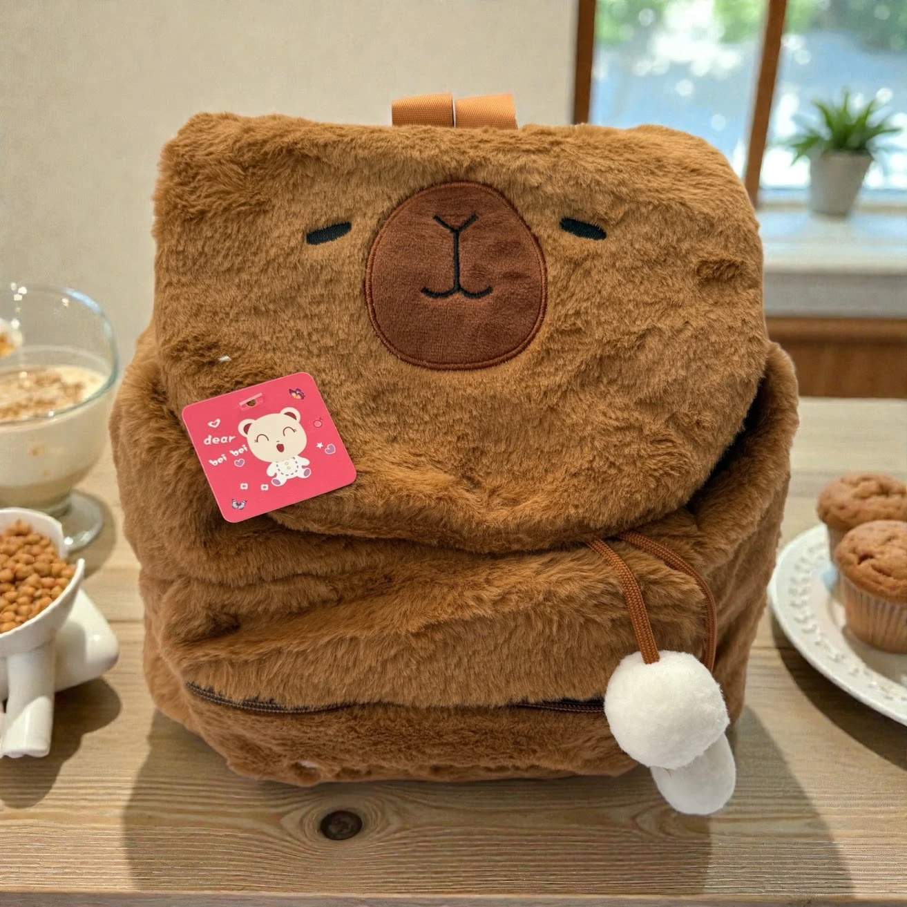 Mochila de felpa marrón con diseño de capibara aplastada, etiqueta rosa visible y pompones blancos en la correa, sobre mesa con desayuno.