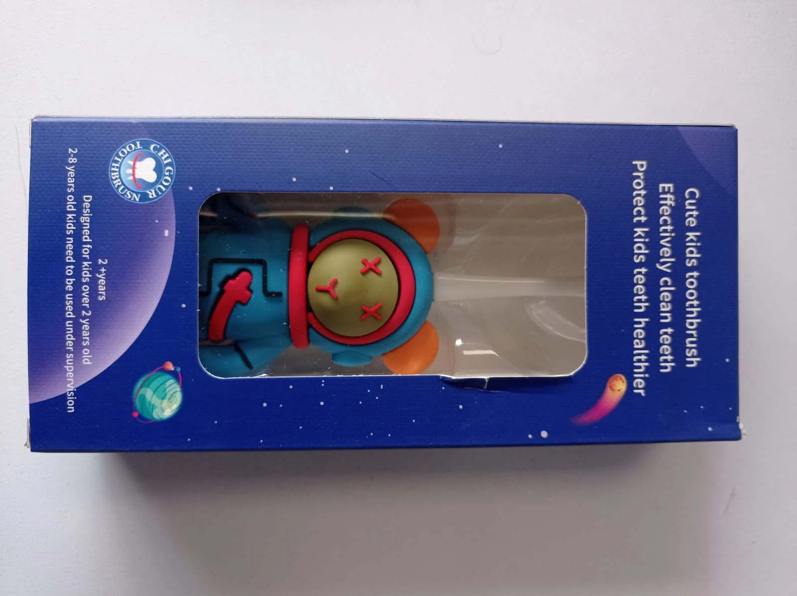 Cepillo dental infantil con diseño de astronauta en caja azul, visible a través de ventana transparente. Incluye cepillo con forma de cohete y detalles en rojo, amarillo y naranja.
