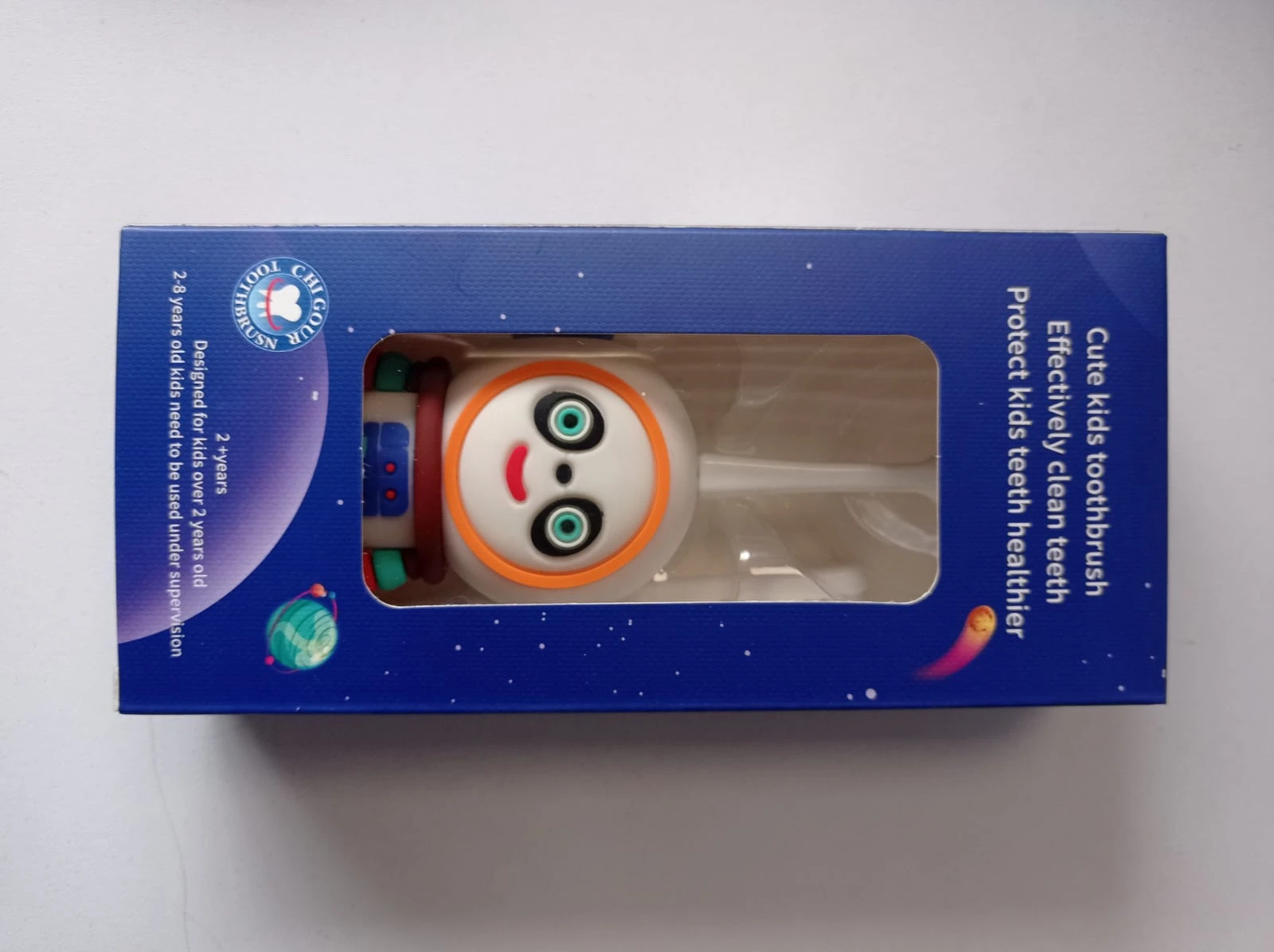 Caja azul con ventana transparente mostrando cepillo infantil con diseño de robot sonriente, ojos verdes y detalles naranjas, para niños de 3 a 6 años.