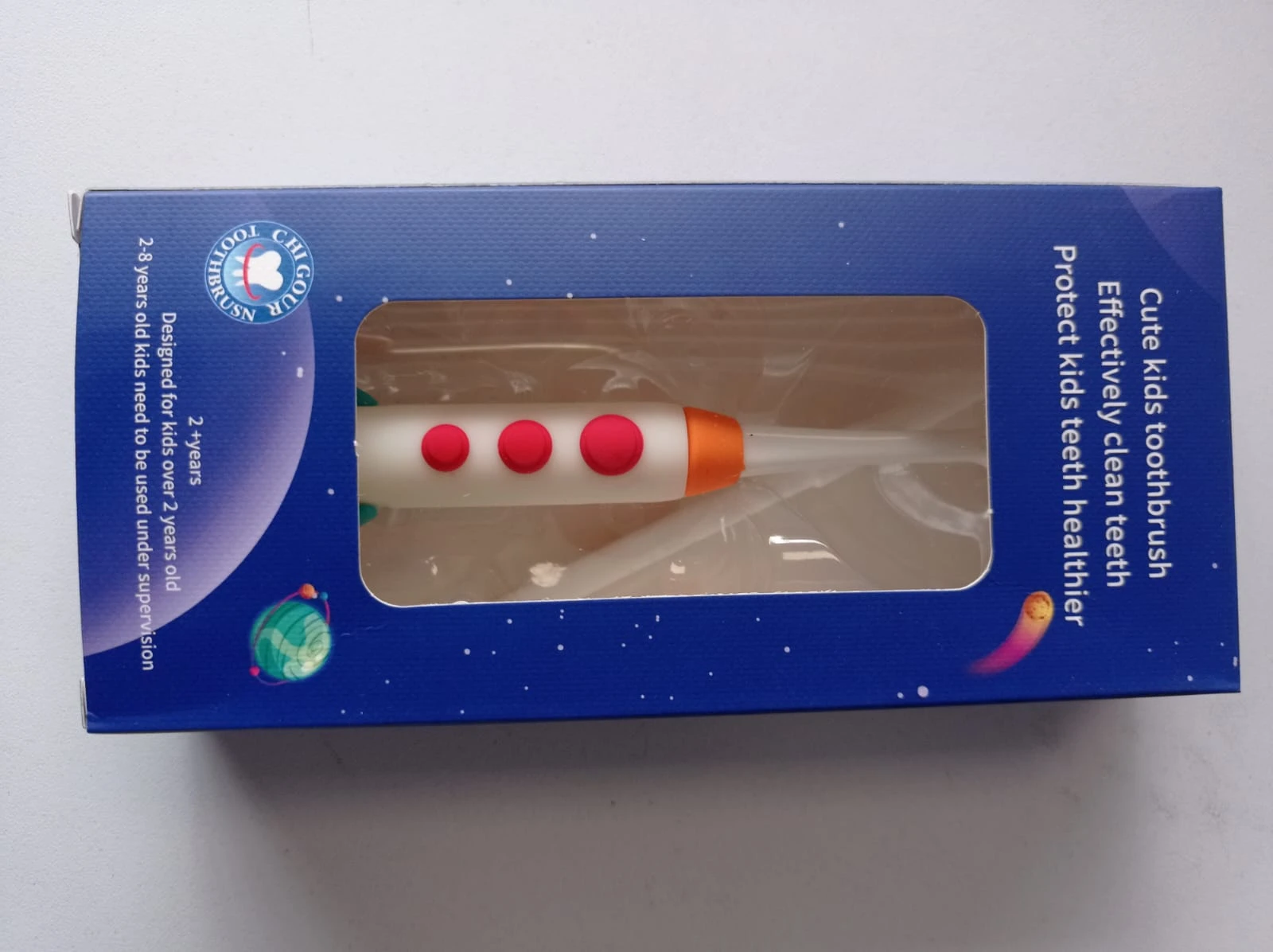 Caja azul de cepillo dental infantil con diseño espacial, incluye cepillo blanco con detalles rojos y naranja, visible a través de ventana transparente.