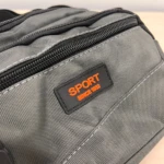 Canguro gris con cremallera negra y etiqueta naranja que dice "SPORT SINCE 1992", mostrando textura de tela resistente y costuras visibles.