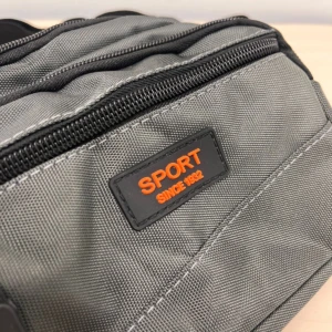 Canguro gris con cremallera negra y etiqueta naranja que dice "SPORT SINCE 1992", mostrando textura de tela resistente y costuras visibles.