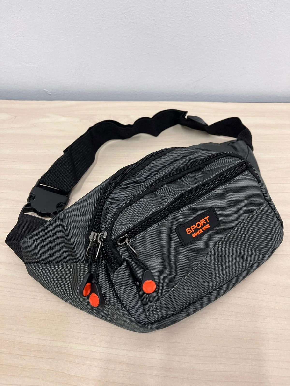 Canguro gris oscuro con tres compartimentos, correa ajustable y detalles en naranja; ideal para llevar activos durante actividades deportivas.