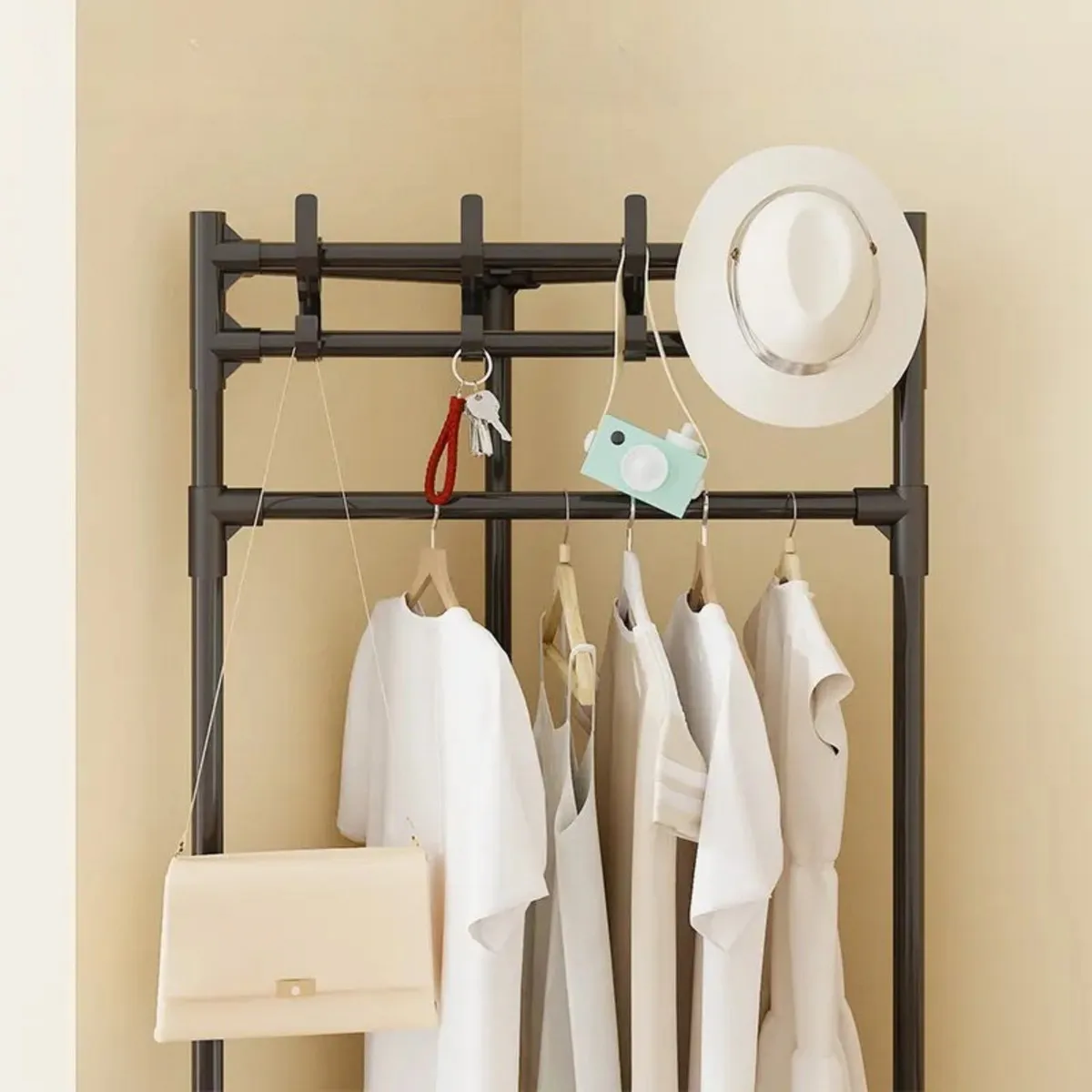 Perchero metálico negro con perchas y gancho superior, exhibiendo ropa blanca, sombrero, bolso y llaves en esquina de pared beige.