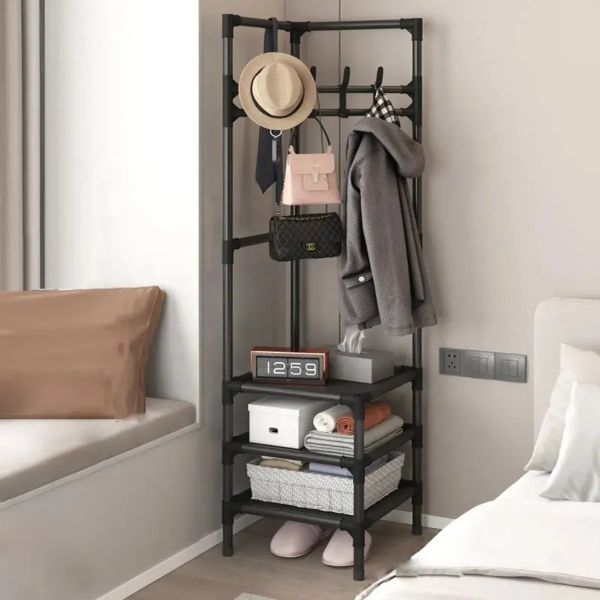 Perchero metálico negro en esquina, con perchas, estantes y cesta; muestra sombrero, bolso, abrigo, reloj digital y calzado. Ideal para organizar espacios reducidos.