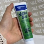 Tubo de pasta dental natural 8 en 1 para dientes sensibles, con etiqueta verde y blanca, sostenido en mano sobre fondo con texto repetido.