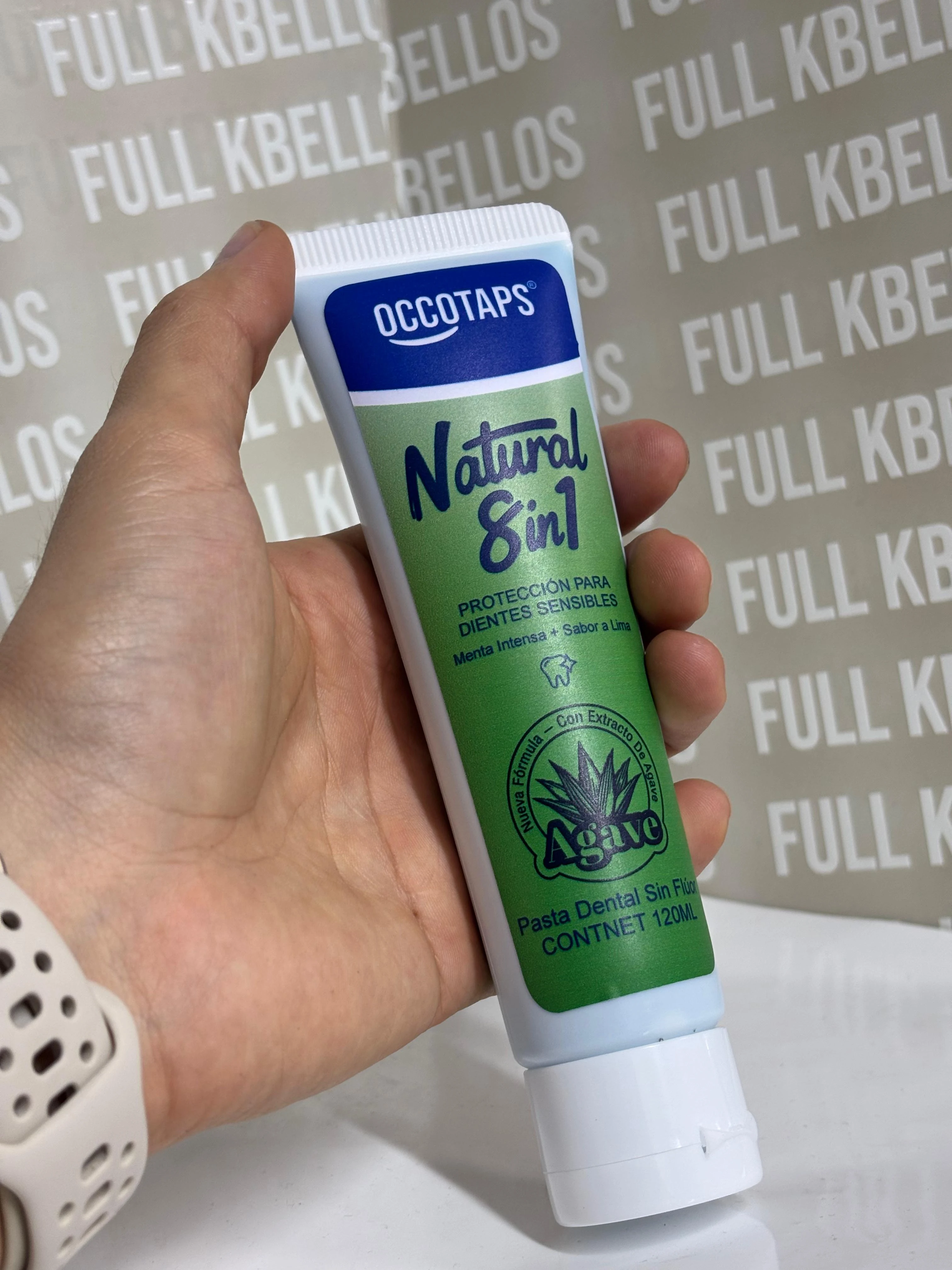Tubo de pasta dental natural 8 en 1 para dientes sensibles, con etiqueta verde y blanca, sostenido en mano sobre fondo con texto repetido.