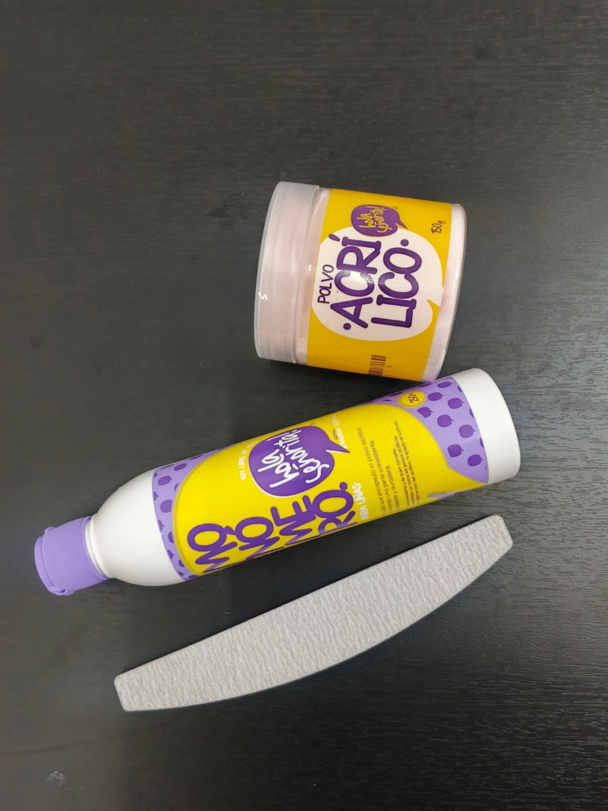 Kit de uñas en acrílico con polvo y monómero, presentados en envases amarillos y morados sobre superficie oscura. Incluye lija blanca para acabado final.