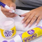 Kit de uñas en acrílico con polvo y monómero. Mano aplicando esmalte sobre uñas naturales, junto a frasco de polvo rosa y botella de monómero violeta sobre toalla blanca.