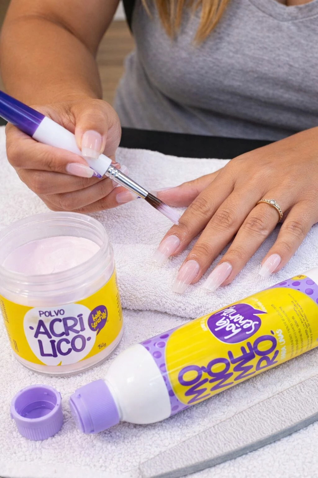 Kit de uñas en acrílico con polvo y monómero. Mano aplicando esmalte sobre uñas naturales, junto a frasco de polvo rosa y botella de monómero violeta sobre toalla blanca.