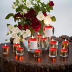 Velas flotantes rojas encendidas en recipientes de vidrio transparente, dispuestas sobre una superficie de madera rústica con flores rojas y blancas al fondo.