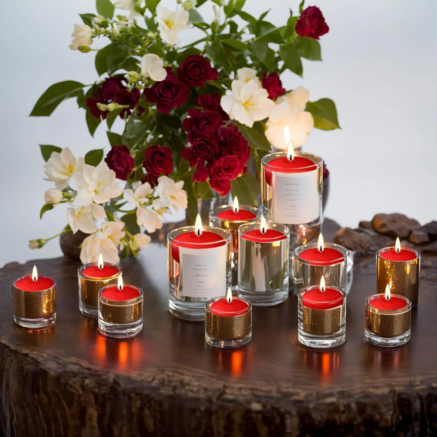 Velas flotantes rojas encendidas en recipientes de vidrio transparente, dispuestas sobre una superficie de madera rústica con flores rojas y blancas al fondo.