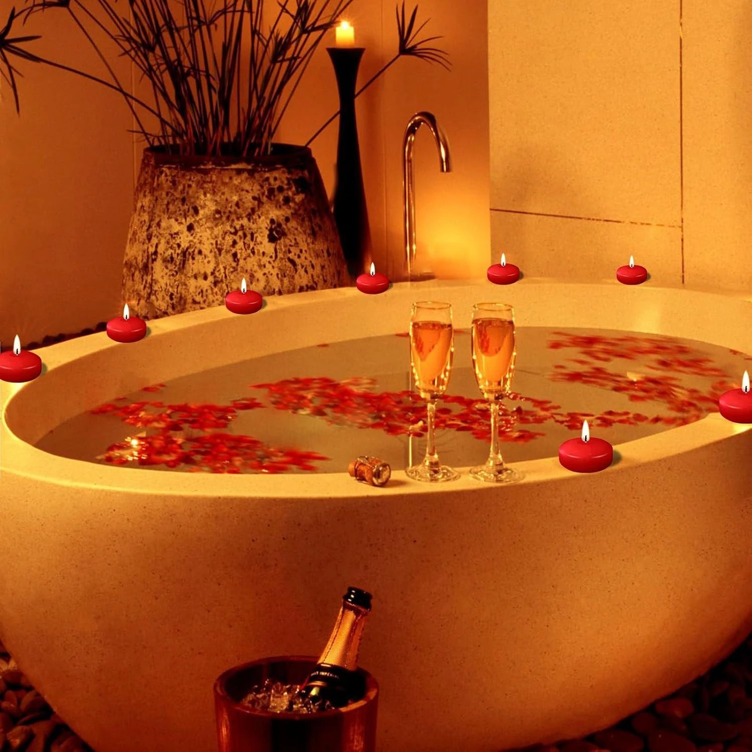 Bañera ovalada beige con velas flotantes rojas, copas de champán y pétalos rosados; ambiente romántico con iluminación cálida y decoración natural.