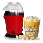 Crispetera de aire compacta roja con tapa transparente, junto a palomitas en taza amarilla y algunas sueltas sobre superficie blanca.
