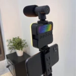 Kit de video con trípode, micrófono y luz LED RGB en color negro, mostrando dispositivo principal con pantalla RGB y micrófono montado superiormente, junto a smartphone fijado en soporte lateral.