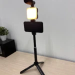 Trípode negro con soporte para celular, luz LED encendida y micrófono montado en la parte superior, sobre superficie de madera clara.