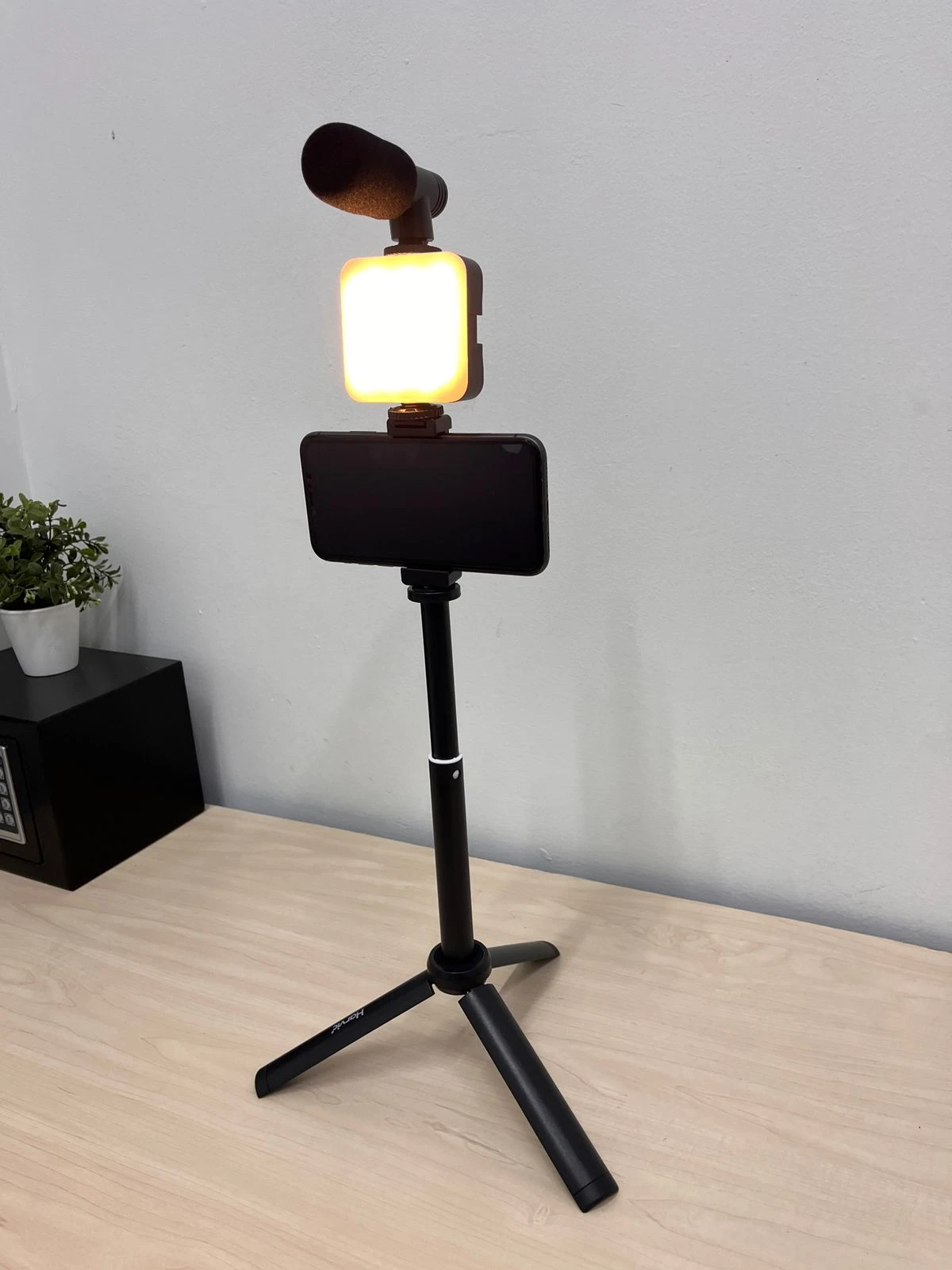 Trípode negro con soporte para celular, luz LED encendida y micrófono montado en la parte superior, sobre superficie de madera clara.