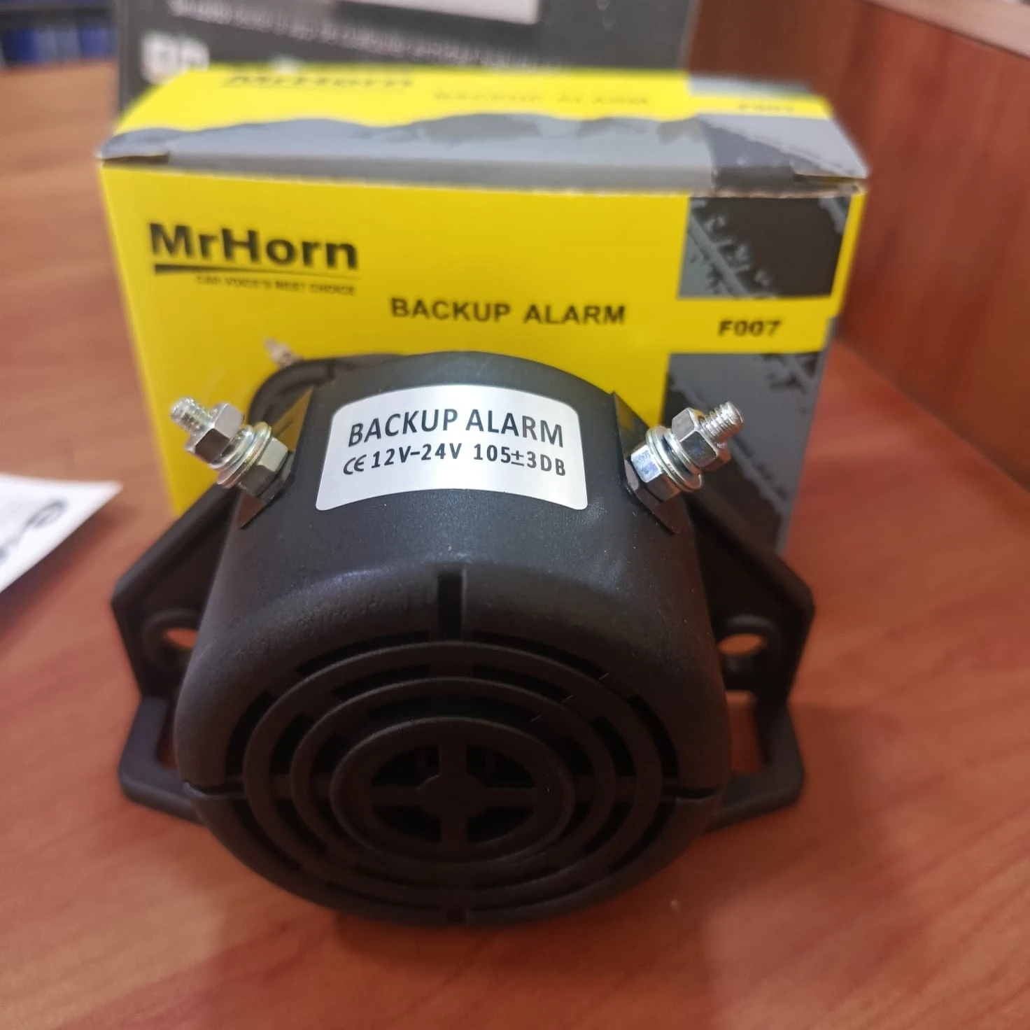 Pito de reversa automático MrHorn, color negro, con etiqueta visible que indica 12V-24V y 105±3dB, montado sobre su base de plástico, acompañado de su caja amarilla en el fondo.