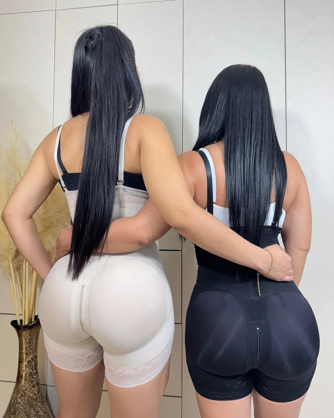 Dos mujeres con fajas Strapple de espalda, una en color crema y otra en negro, mostrando la cremallera frontal y ajuste en cintura y cadera.