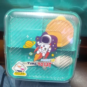 Lonchera rectangular de plástico verde con tapa transparente, diseño espacial con astronauta y planetas, incluye utensilios blancos y naranja visibles.