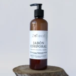 Bote de jabón corporal natural Zoieco en vidrio ámbar con dispensador negro, sobre base de madera rústica. Etiqueta blanca con texto negro y detalles florales.