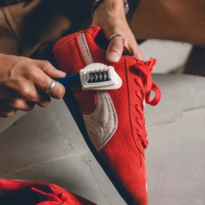Puma Suede en rojo siendo limpiada con cepillo de cerdas blancas y mango azul, mostrando textura de gamuza y cordones rojos.