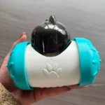 Dispensador interactivo para perros en color blanco con detalles en azul turquesa, forma de cápsula con empuñaduras laterales y logotipo de huella de pata en la parte frontal.