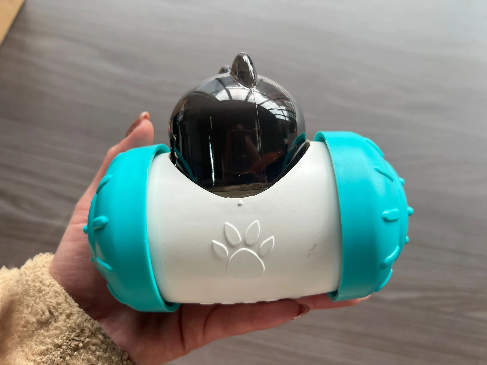 Dispensador interactivo para perros en color blanco con detalles en azul turquesa, forma de cápsula con empuñaduras laterales y logotipo de huella de pata en la parte frontal.