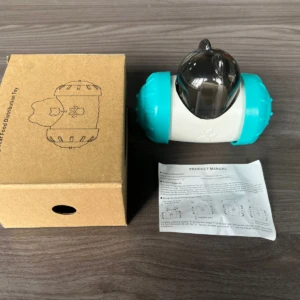 Dispensador interactivo para perros en color turquesa y blanco, con caja de cartón marrón y manual incluido, sobre superficie de madera.