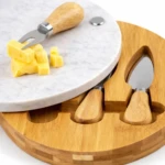 Tabla de quesos redonda con superficie de mármol blanco y base de bambú natural, incluye cuchillo de servir y dos utensilios de madera con mango, sobre fondo blanco.