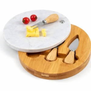 Tabla redonda de quesos con superficie de mármol blanco y base de bambú, incluye dos utensilios de madera y queso cortado con tomates cherry encima.