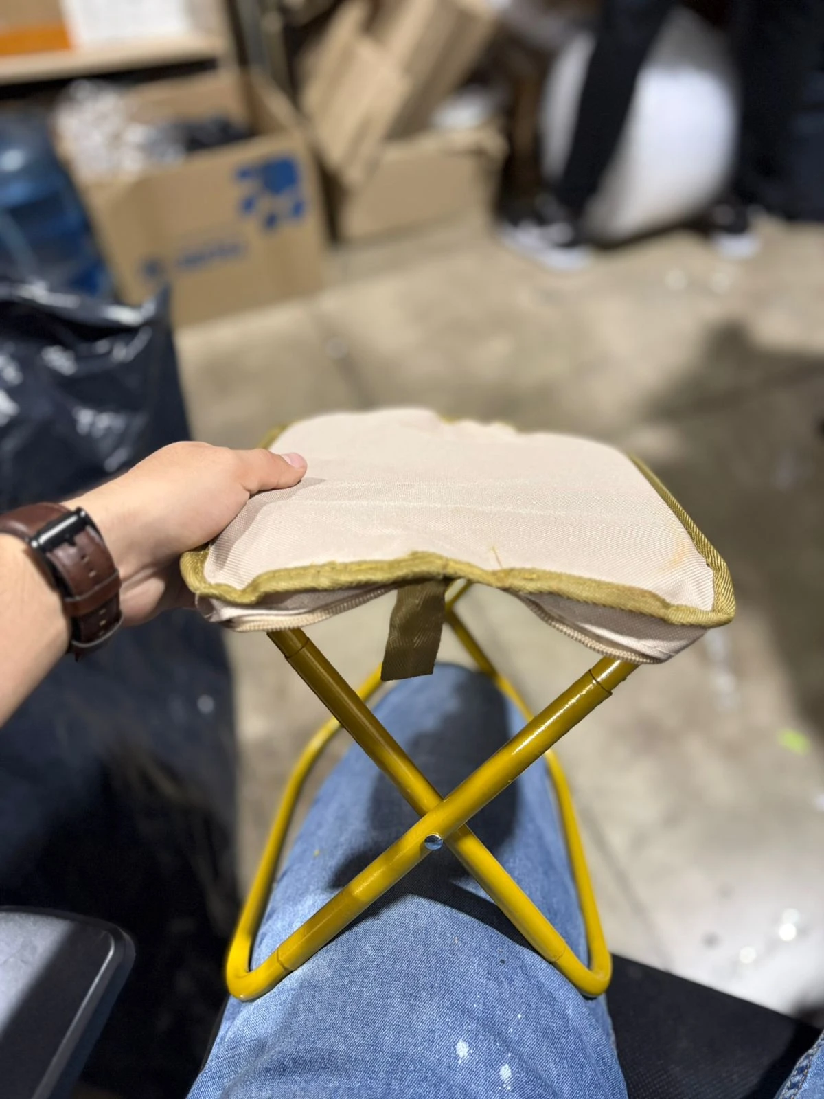 Silla plegable de acero amarillo con asiento acolchado beige, sostenida por mano con reloj de cuero, sobre rodilla con jeans.