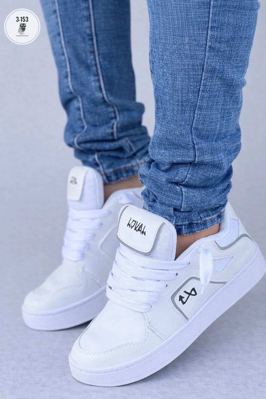 Zapatilla deportiva blanca de Lokal Colombia con detalles en gris, vista frontal de perfil, usada con jeans azul. Suela de goma visible.